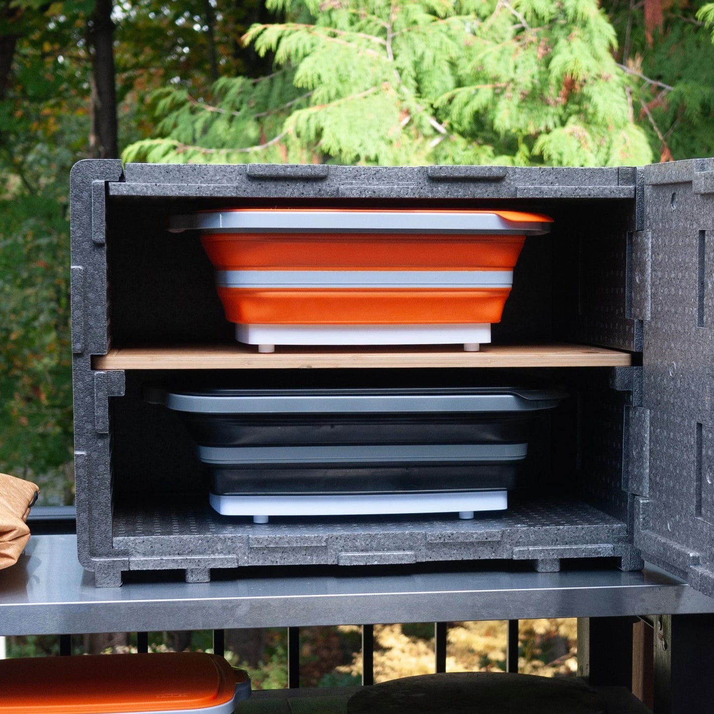 A 'Que Crib' meat resting station open to show its interior, with shelves holding orange and black containers. The unit appears to be made of gray material and is located outdoors with trees in the background.