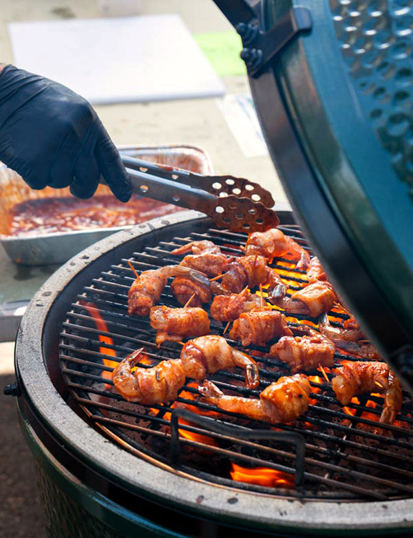 Grilled shrimp skewers cooking on a charcoal grill with a person wearing a black glove using metal tongs to turn them, with the grill lid partially open in the foreground.