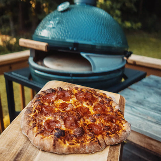 Pepperoni pizza on a wooden board with a blue ceramic pizza oven in the background.